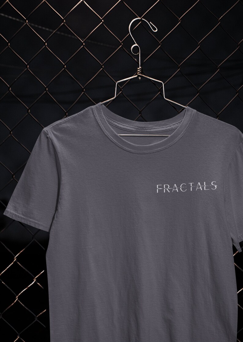 May include: A gray t-shirt with the word "FRACTALS" printed in white on the front. The shirt is hanging on a metal hanger against a chain link fence.