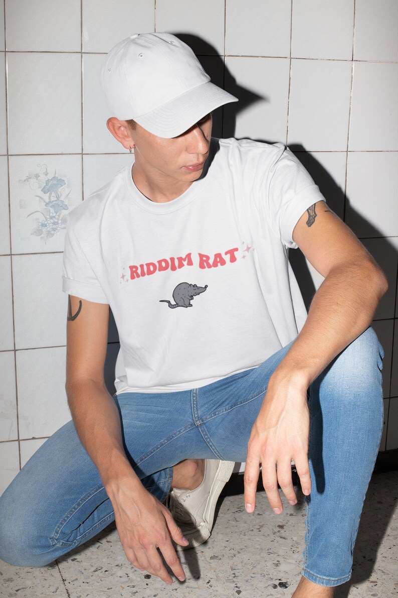May include: A white t-shirt with a cartoon rat and the text "RIDDIM RAT" in pink lettering. The person is wearing blue jeans and a white baseball cap.