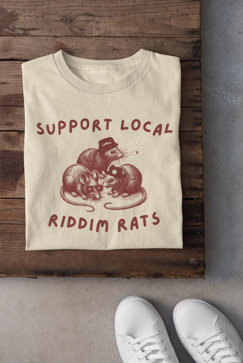 Pu&ograve; includere: Una t-shirt beige con il testo "SUPPORT LOCAL" e "RIDDIM RATS" stampato in marrone. Il design presenta tre ratti di cartoni animati con cappelli e occhiali da sole.
