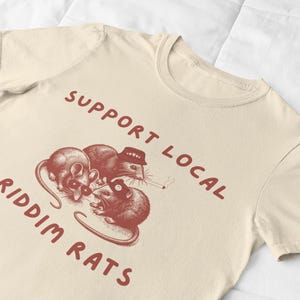 Pu&ograve; includere: Una maglietta beige con una grafica rossa di tre ratti che indossano cappelli e tengono un giradischi. Il testo "Support Local Riddim Rats" &egrave; stampato in rosso sulla maglietta.