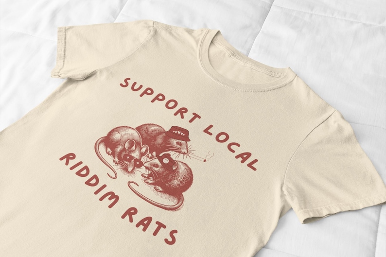 Pu&ograve; includere: Una maglietta beige con una grafica rossa di tre ratti che indossano cappelli e tengono un giradischi. Il testo "Support Local Riddim Rats" &egrave; stampato in rosso sulla maglietta.