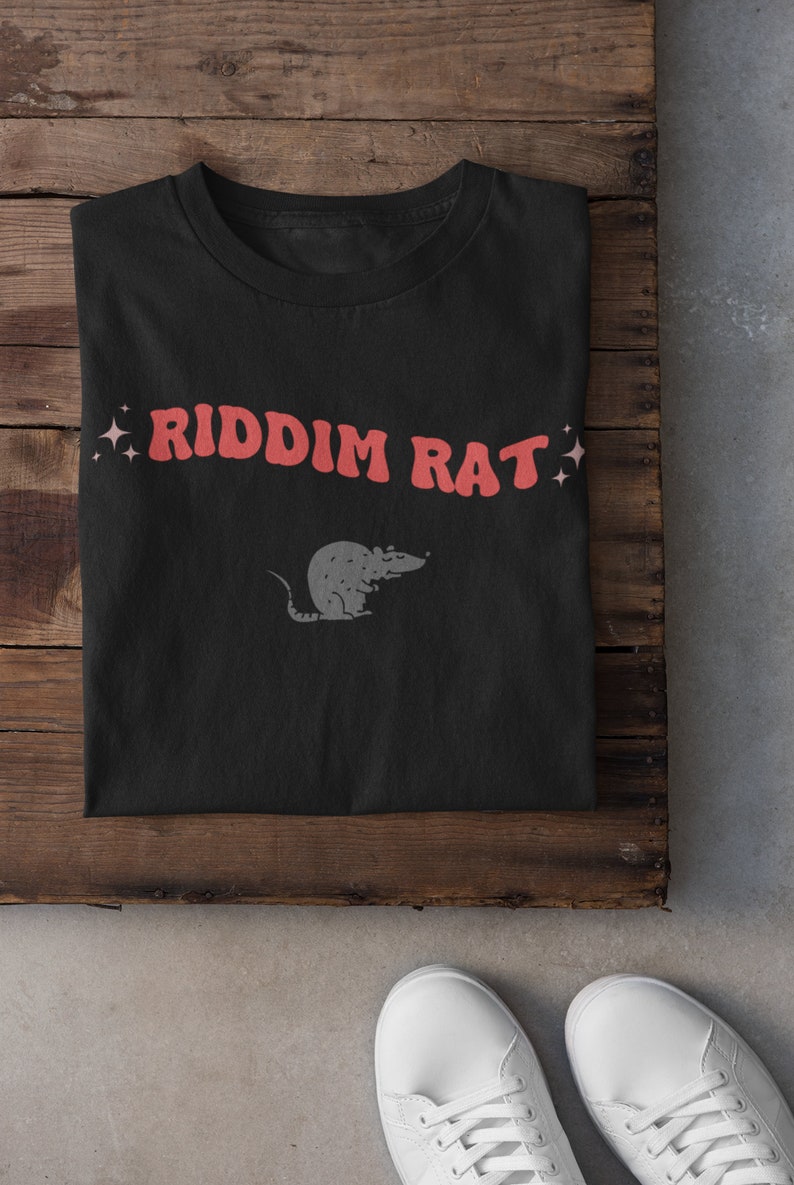 May include: Black t-shirt with the text "RIDDIM RAT" in red letters and a small gray rat illustration. The shirt is folded and laid on a wooden surface.