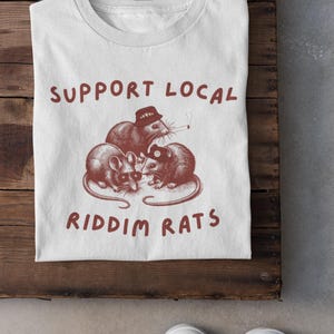 Pu&ograve; includere: Maglietta bianca con una grafica rossa di tre ratti che indossano cappelli e occhiali da sole. Il testo "SUPPORT LOCAL RIDDIM RATS" &egrave; stampato in rosso sulla maglietta.