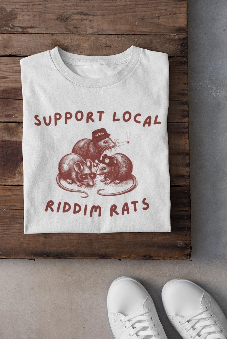 Pu&ograve; includere: Maglietta bianca con una grafica rossa di tre ratti che indossano cappelli e occhiali da sole. Il testo "SUPPORT LOCAL RIDDIM RATS" &egrave; stampato in rosso sulla maglietta.