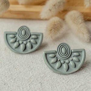 Puede incluir: Un par de pendientes grises en forma de semicírculo con un diseño detallado que presenta un círculo con un patrón de espiral y formas de hojas alrededor del borde.