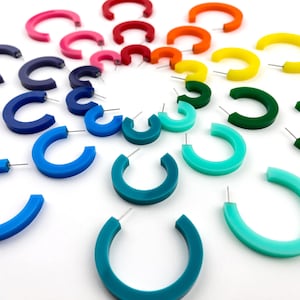 Puede incluir: Un conjunto de pendientes de aro coloridos en un degradado arcoíris. Los pendientes están hechos de un plástico grueso y liso y están dispuestos en círculo sobre un fondo blanco.