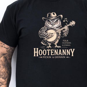 May include: Black t-shirt with a graphic of an owl playing a banjo. The text on the shirt reads "Folk Feathers & Strings" and "Hootenanny - Pickin' & Grinnin' - "