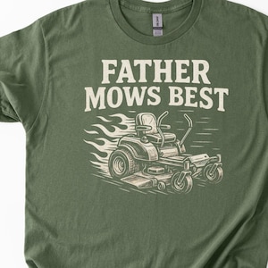 May include: An olive green t-shirt with the text "FATHER MOWS BEST" above a graphic of a riding lawn mower. The mower is depicted in black and white with flames and speed lines. White sneakers are visible in the bottom right corner.