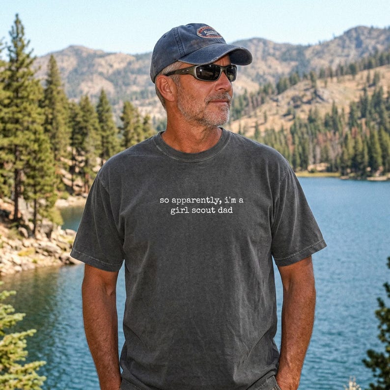 May include: A charcoal gray t-shirt with the text "so apparently, I'm a girl scout dad" in white. The man in the image is wearing a blue baseball cap and sunglasses. The background features a lake and mountains.