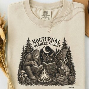 May include: An ivory t-shirt featuring a graphic print of a Bigfoot, a moth, and a jackalope reading books around a campfire in a forest. The text reads "NOCTURNAL READERS SOCIETY" and "NATURAL HISTORIES OF THE OBSCURE & UNSEEN" on the book cover.