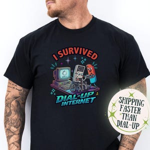 May include: Black t-shirt with a colorful graphic that reads "I Survived Dial-Up Internet." The design features a retro computer, a game controller in a wheelchair, and a pill character. The shirt has a vintage, nostalgic feel.