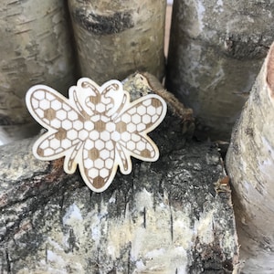 May include: A wooden bee cutout with a honeycomb pattern. The bee is light brown and has a white honeycomb pattern. The bee is sitting on a piece of birch bark.