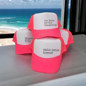 Puede incluir: Una colecci&oacute;n de gorras de camionero de color rosa ne&oacute;n y blanco con partes traseras de malla y paneles frontales de espuma. Las gorras muestran texto impreso en negro, incluyendo las frases I'm Here for the Gangbang, Mayor Titty y same penis forever.