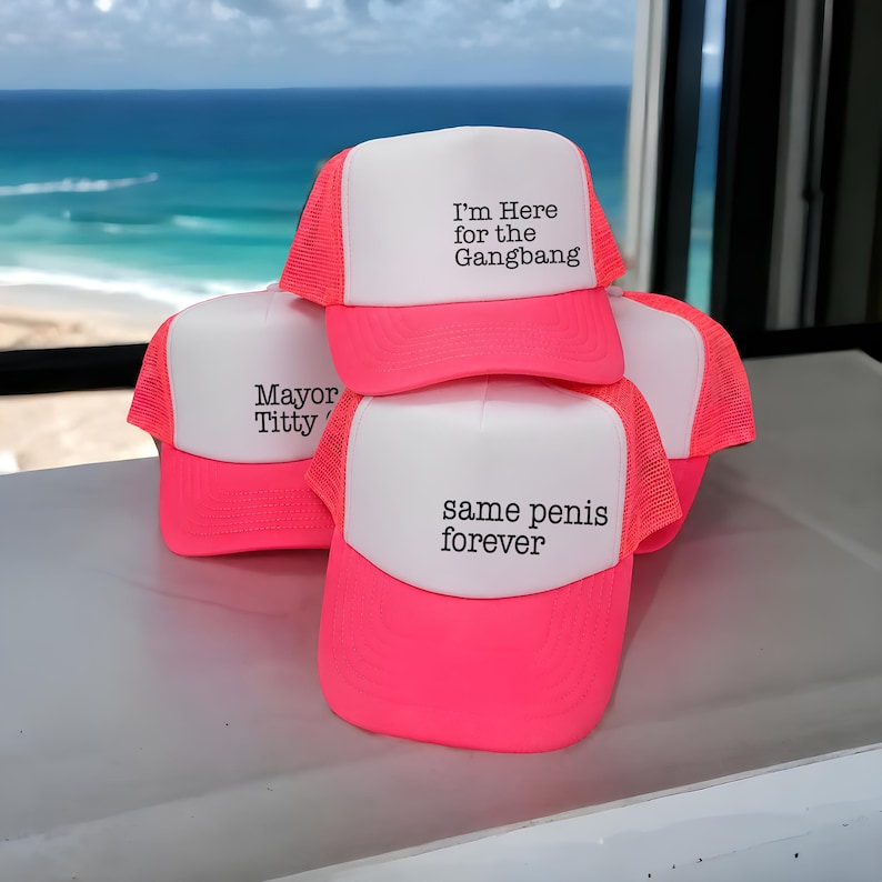 Puede incluir: Una colecci&oacute;n de gorras de camionero de color rosa ne&oacute;n y blanco con partes traseras de malla y paneles frontales de espuma. Las gorras muestran texto impreso en negro, incluyendo las frases I'm Here for the Gangbang, Mayor Titty y same penis forever.
