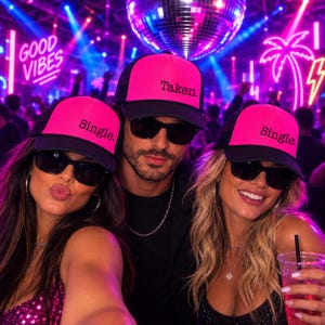 Puede incluir: Tres gorras de camionero de color rosa ne&oacute;n y negro con el texto 'Single.' en dos gorras y 'Taken.' en la gorra central. Cada gorra tiene una parte trasera de malla y un panel frontal s&oacute;lido. Las gorras son usadas en una discoteca.