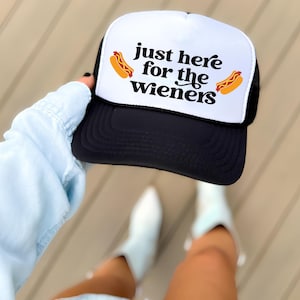 May include: Black and white trucker hat with the text "just here for the wieners" and two cartoon hot dogs. The hat has a white front panel and a black mesh back.