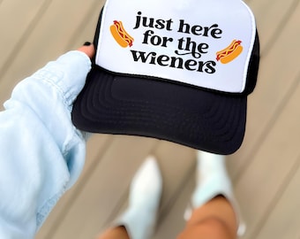 May include: Black and white trucker hat with the text "just here for the wieners" and two cartoon hot dogs. The hat has a white front panel and a black mesh back.