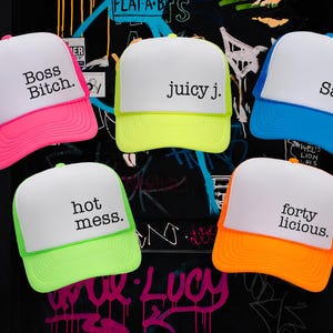 Puede incluir: Cinco gorras de camionero de espuma con paneles frontales blancos y viseras y mallas traseras de colores ne&oacute;n. Las gorras presentan texto en negro: Boss Bitch, juicy j., Salty, hot mess. y forty licious. Los colores incluyen rosa, amarillo, azul y naranja.