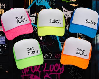 Könnte beinhalten: Fünf Schaumstoff-Trucker-Caps mit weißen Vorderteilen sowie neonfarbenen Schirmen und Netzeinsätzen. Die Caps tragen schwarzen Text: Boss Bitch, juicy j., Salty, hot mess. und forty licious. Die Farben umfassen Neonpink, Neongelb, Blau und Orange.