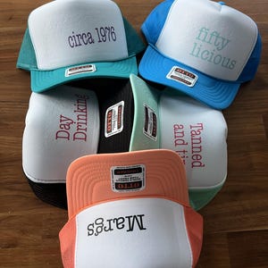 Puede incluir: Una colecci&oacute;n de cinco gorras de camionero con frente de espuma en varios colores, incluyendo verde azulado, azul, negro, menta y melocot&oacute;n. Cada gorra presenta un panel frontal blanco con texto impreso: circa 1976, fifty licious, Day Drinking, Tanned and ti y Margs.