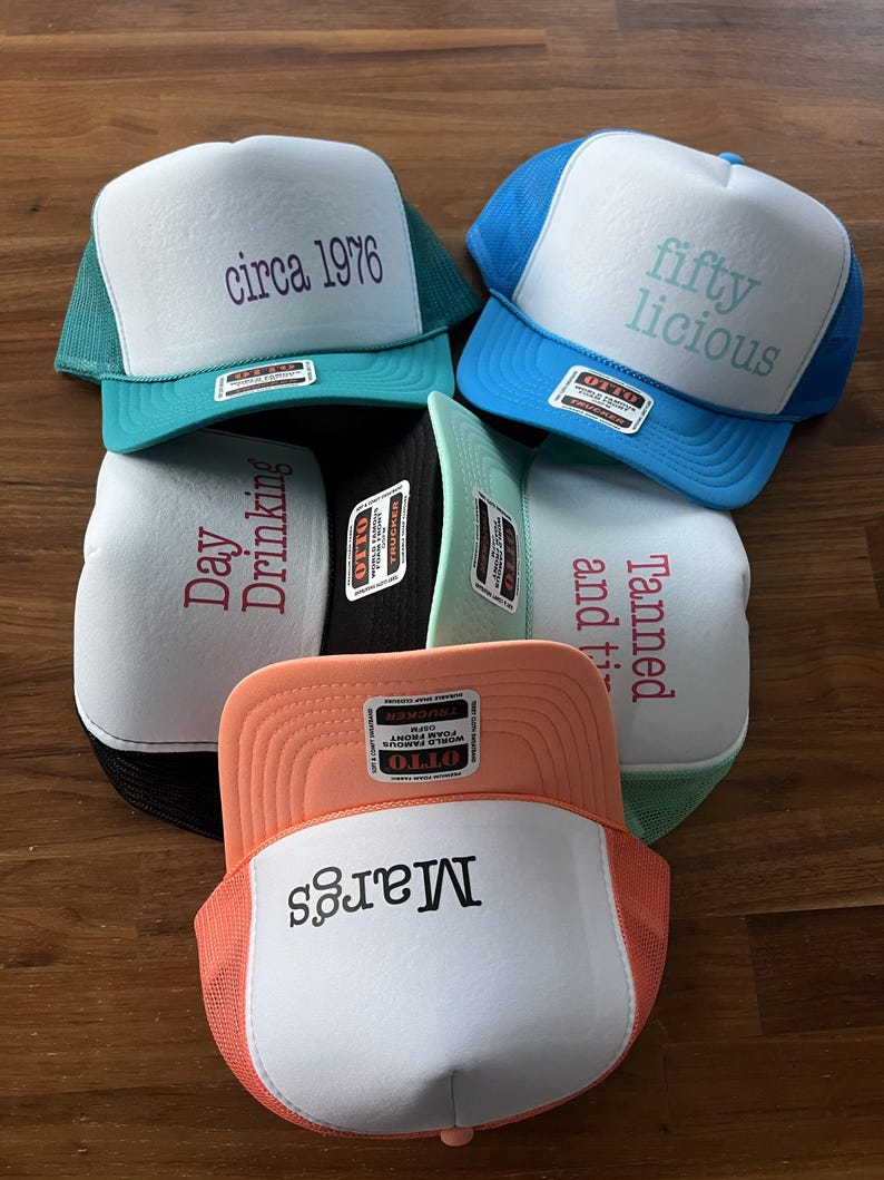 Puede incluir: Una colecci&oacute;n de cinco gorras de camionero con frente de espuma en varios colores, incluyendo verde azulado, azul, negro, menta y melocot&oacute;n. Cada gorra presenta un panel frontal blanco con texto impreso: circa 1976, fifty licious, Day Drinking, Tanned and ti y Margs.
