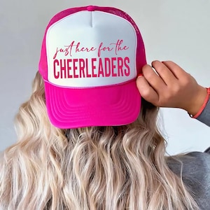 Puede incluir: Una gorra tipo camionero de color rosa brillante y blanco con parte trasera de malla y panel frontal sólido. El panel frontal blanco muestra el texto "just here for the CHEERLEADERS" en una fuente rosa vibrante, con el texto en cursiva.