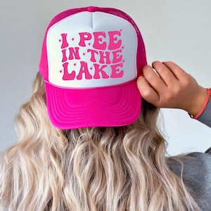 a woman wearing a pink and white trucker hat with i pee in the lake