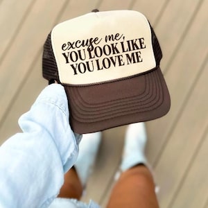 May include: A brown and white trucker hat with the text "excuse me, you look like you love me" in black. The hat has a mesh back and a curved brim. The hat is being held up.