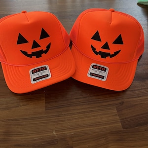 May include: Two bright orange trucker hats featuring a black jack-o'-lantern face graphic on the front foam panel. Each hat includes a white sticker on the brim with the text OTTO WORLD FAMOUS FOAM FRONT OSFM TRUCKER and a durable snap closure.