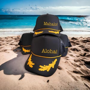 Puede incluir: Una colecci&oacute;n de gorras tipo camionero negras con paneles traseros de malla y bordados amarillos. Los paneles frontales muestran el texto Mahalo! o Aloha! en amarillo, junto con patrones de hojas bordados en el borde de la visera.