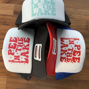 Puede incluir: Varios gorros de camionero con la frase "I PEE IN THE LAKE" impresa en la parte delantera. Los gorros tienen un panel frontal blanco con el texto en varios colores y una parte trasera de malla en negro, rojo y azul.