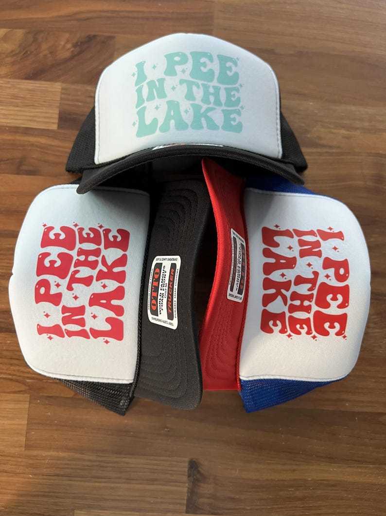 Puede incluir: Varios gorros de camionero con la frase "I PEE IN THE LAKE" impresa en la parte delantera. Los gorros tienen un panel frontal blanco con el texto en varios colores y una parte trasera de malla en negro, rojo y azul.