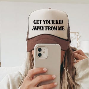 May include: A white and brown trucker hat with the text "GET YOUR KID AWAY FROM ME" in black. The hat has a brown brim and mesh back. The image shows a person holding a phone.