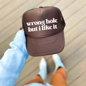 May include: Brown trucker hat with the text "wrong hole but i like it" in white block letters. The hat has a mesh back and a curved brim. A brown rope detail is on the front of the hat.