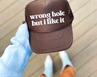 May include: Brown trucker hat with the text "wrong hole but i like it" in white block letters. The hat has a mesh back and a curved brim. A brown rope detail is on the front of the hat.