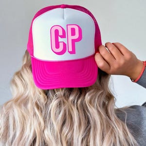 May include: A hot pink trucker hat with a white front panel featuring the letters "CP" in a layered pink design. The hat has a mesh back and a curved brim. The image shows the back of a person wearing the hat.