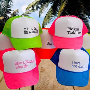 Puede incluir: Una colecci&oacute;n de gorras de camionero de colores ne&oacute;n con paneles frontales blancos. Las gorras presentan varias frases impresas en texto negro, rosa o azul, incluyendo Y.O.L.O. BE A HOE, Pickle Tickler, Save a Horse Ride Me, i love hot dads y Sava.