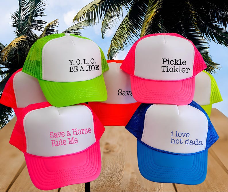 Puede incluir: Una colecci&oacute;n de gorras de camionero de colores ne&oacute;n con paneles frontales blancos. Las gorras presentan varias frases impresas en texto negro, rosa o azul, incluyendo Y.O.L.O. BE A HOE, Pickle Tickler, Save a Horse Ride Me, i love hot dads y Sava.