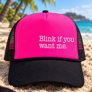 Puede incluir: Una gorra tipo trucker de color rosa brillante con visera negra y paneles traseros de malla negra. El panel frontal presenta el texto blanco "Blink if you want me.". La gorra tiene un frente de espuma estructurado y un bot&oacute;n negro.