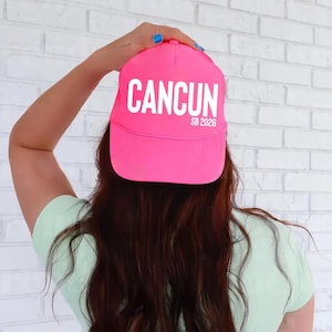May include: A bright pink baseball cap with the word "CANCUN" in white block letters. Below the word "CANCUN" is the text "SB 2026" in white. The cap is being held up by a person.