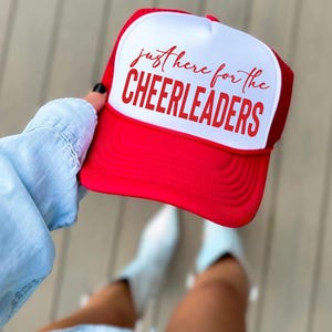 May include: A red and white trucker hat featuring a white front panel with the red text "just here for the CHEERLEADERS" printed in a combination of script and bold block lettering. The hat has a solid red brim and a red mesh back panel.