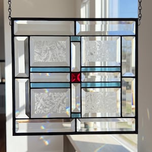May include: A square stained glass window hanging featuring a grid of beveled clear glass panels, textured glass inserts, horizontal light blue glass strips, and a single central square red glass accent, all held together by black metal cames.