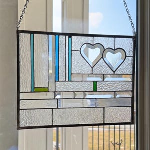 May include: A rectangular stained glass suncatcher featuring a geometric arrangement of clear textured glass panels. The design includes two heart-shaped cutouts, vertical blue and green glass accents, and a dark metal frame with a hanging chain.