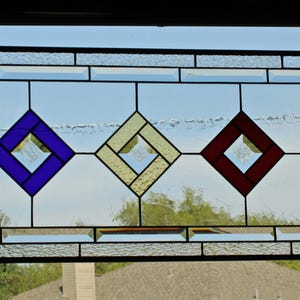 May include: A rectangular stained glass panel featuring a black metal frame. The design includes three central diamond shapes in purple, cream, and red, surrounded by clear textured glass panes and a geometric border pattern.