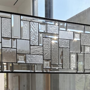 May include: A rectangular stained glass panel featuring a geometric arrangement of clear glass pieces with varied textures, including smooth, rippled, and pebbled surfaces, all encased within a thin black metal frame and suspended by metal chains.