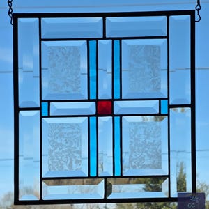 May include: A stained glass window with a geometric design. The window features clear, light blue, and red glass panes. The design includes rectangular and square shapes, with a central red square and blue vertical stripes. The glass has a textured, frosted appearance.