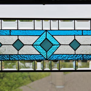 May include: A stained glass window panel with a geometric design featuring blue and white glass. The panel is framed in black and has a hanging chain.