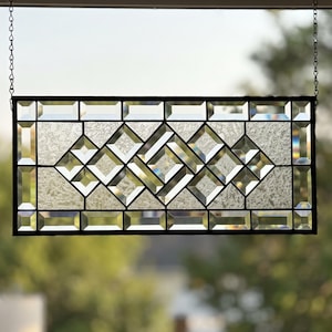 May include: A rectangular stained glass window hanging featuring a geometric pattern of clear beveled glass pieces and textured glass panels. The design is framed in a dark metal border and suspended by two silver chains from the top frame.