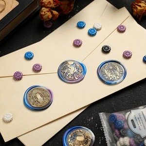 May include: A close-up of a variety of wax seals with different designs. The seals are on a beige surface with a variety of small decorative flowers. The seals are blue, purple, and gold.