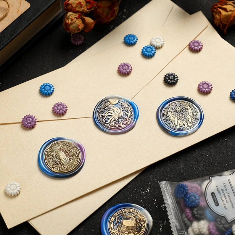 May include: A close-up of a variety of wax seals with different designs. The seals are on a beige surface with a variety of small decorative flowers. The seals are blue, purple, and gold.